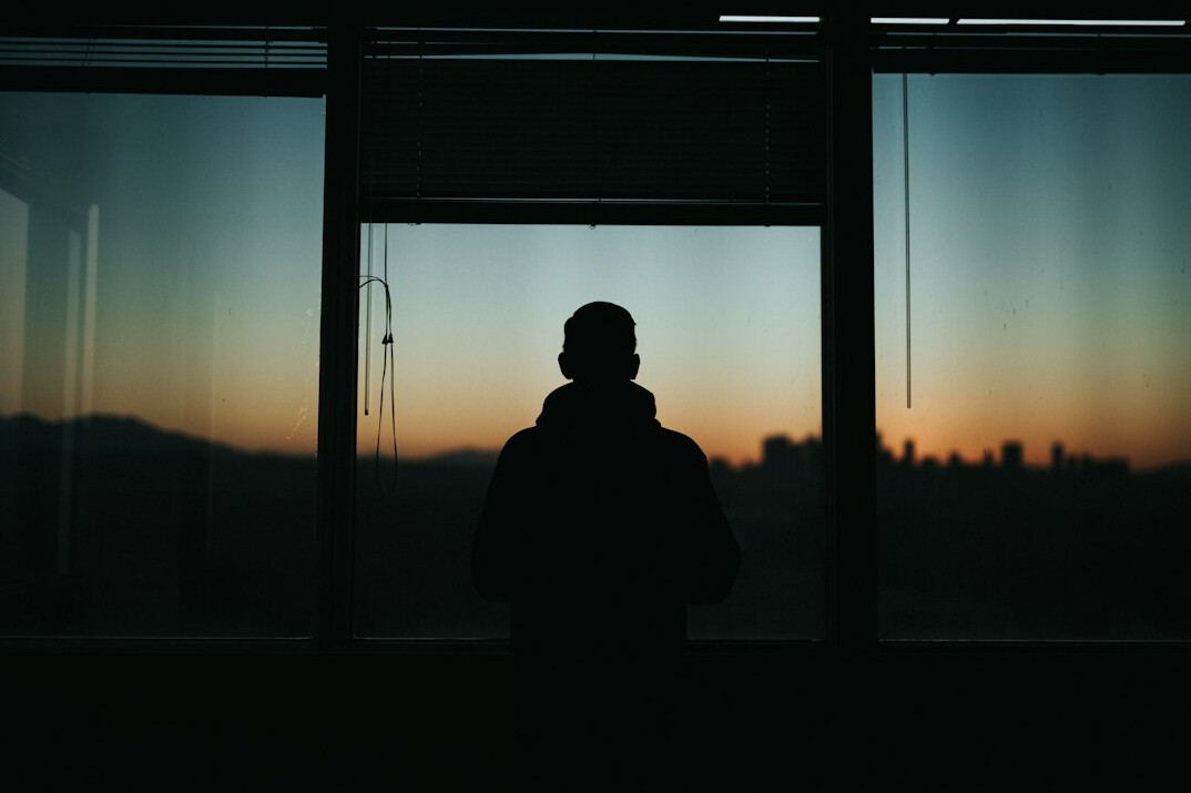 Silhouette of a person standing alone facing a large glowing window at dusk